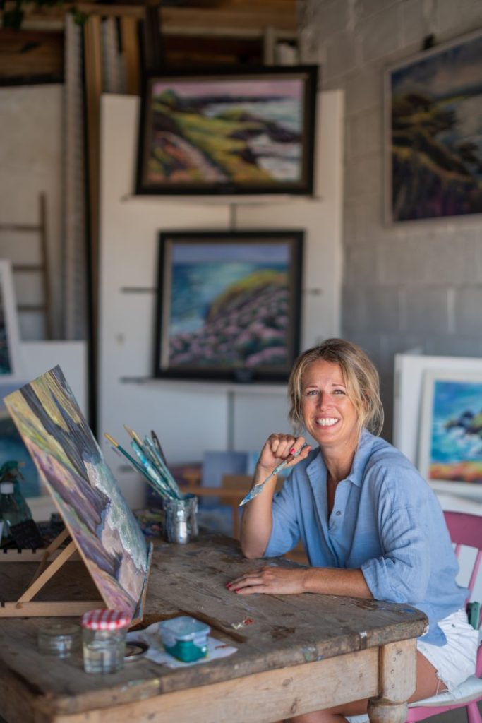 Bobby Magee sitting at a desk in her studio, holding a paintbrush with a painting in front of her.
Photo: Jim Wileman